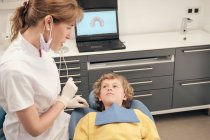 Mujer en uniforme médico hablando con un pequeño paciente en el consultorio del dentista - foto de stock