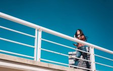 Desde abajo mujer joven confiada con teléfono inteligente y auriculares caminando en el puente en el fondo del cielo azul - foto de stock