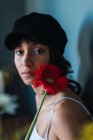 Молодая очаровательная брюнетка в кепке смотрит в камеру и держит свежий красный цветок в комнате — стоковое фото