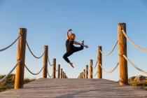 Young ballerina in black wear with upped leg in air on footbridge and blue sky in sunny day — Stock Photo
