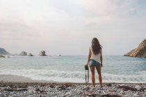Vue arrière de la femme en short haut et en denim avec appareil photo debout et regardant la mer sur la côte près des rochers — Photo de stock