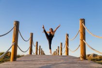 Молодая балерина в черной одежде с поднятой ногой в воздухе на пешеходном мосту и голубым небом в солнечный день — стоковое фото