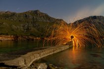 Longa exposição de silhueta de humano com queima redonda e fogos de artifício em cais perto de água e montanhas à noite — Fotografia de Stock