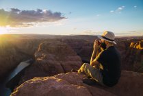 Vue latérale du gars en chapeau profitant d'une boisson chaude et regardant coucher le soleil tout en étant assis près de merveilleux canyon sur la côte ouest des États-Unis — Photo de stock