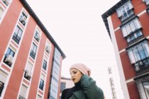 D'en bas de femme confiante avec le maquillage et la tête couverte posant sur la rue contre les bâtiments rouges de la ville — Photo de stock