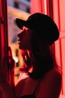 Вид збоку молодої стрункої жінки в кепці, що стоїть біля балкона в кімнаті в почервоніння вночі — стокове фото