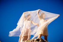 Desde abajo mujeres misteriosas jóvenes con las manos levantadas sosteniendo textiles blancos y posando sobre rocas y cielo azul - foto de stock
