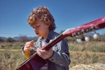 Cute boy playing acoustic guitar and singing while sitting in huge roll of dry hay on sunny day in countryside — Stock Photo