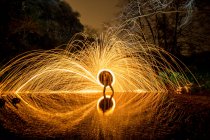Longue exposition de silhouette humaine avec feu rond et feux d'artifice sur les rochers près de l'eau dans la soirée — Photo de stock