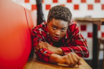 Mujer afroamericana confiada acostada en asientos en la cafetería y mirando a la cámara - foto de stock