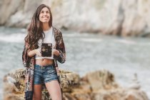 Smiling young woman holding camera on sea coast — Stock Photo
