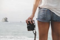 Back view of woman with retro photo camera standing on beach and looking at sea view — Stock Photo