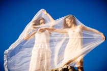 From below young mysterious women with upped hands holding white textile and posing on rocks and blue sky — Stock Photo