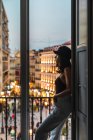 Молодая мечтательная женщина в кепке стоит на балконе на улице с огнями в вечернее время — стоковое фото