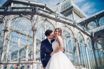 Снизу молодой элегантный мужчина обнимает женщину в свадебном платье возле ретро дворца со множеством окон в солнечный день — стоковое фото