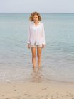 Jeune femme charmante en chemise blanche marchant dans l'eau de mer et regardant la caméra — Photo de stock