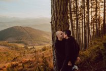 Cheerful homosexual couple kissing near tree in forest and picturesque view of valley — Stock Photo