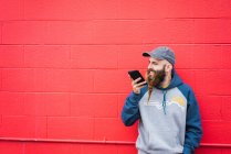 Tipo atractivo con barba trenzada hablando en el teléfono inteligente mientras se inclina pared roja en la calle de la ciudad - foto de stock
