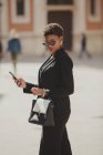 Уверенная афроамериканская элегантная женщина в костюмах и солнцезащитных очках держит сумку и мобильный телефон на улице — стоковое фото