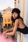 Junge schlanke Frau mit Mütze blickt in die Kamera und steht auf dem Balkon auf der Straße — Stockfoto