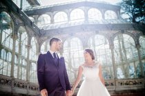 Von unten hält ein junger eleganter Mann eine Frau im Hochzeitskleid in der Nähe des Retro-Palastes mit vielen Fenstern an sonnigen Tagen an den Händen — Stockfoto