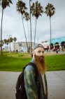 Вид збоку бородатого хлопця з рюкзаком, який дивиться на камеру, стоячи на тротуарі на вулиці сучасного міста — стокове фото
