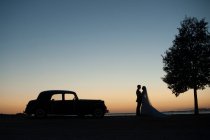 Seitenansicht der Silhouetten von Frischvermählten Händchen haltend in der Nähe von Retro-Automobil am Ufer und blauem Himmel am Abend — Stockfoto