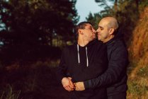 Homosexual couple embracing and kissing on walkway in forest in sunny day on blurred background — Stock Photo