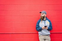Attraktiver Kerl mit geflochtenem Bart surft im Smartphone, während er sich an eine rote Wand auf der Straße lehnt — Stockfoto