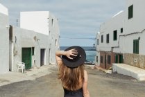 Back view of stylish woman touching hat while standing on shabby street of small coastal town on cloudy day near sea — Stock Photo