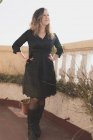 Junge elegante, gut gelaunte Dame im Kleid mit der Hand auf der Hüfte, Brille haltend und bei sonnigem Wetter auf dem Balkon posierend — Stockfoto
