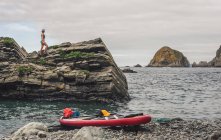 Woman in swimwear standing on rough cliff near red paddle board and waving sea in a stony shore — Stock Photo