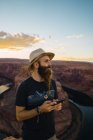 Beau homme avec téléphone portable tout en se tenant contre le magnifique canyon et la rivière pendant le coucher du soleil sur la côte ouest des États-Unis — Photo de stock