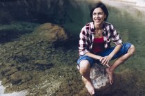 Feliz mujer adulta sentada en la roca en el tranquilo agua transparente del lago disfrutando de la naturaleza y sonriendo - foto de stock