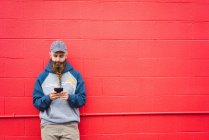 Привлекательный парень с плетеной бородой просматривает смартфон, опираясь на красную стену на городской улице — стоковое фото