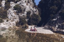 Vue latérale du jeune couple assis sur la côte rocheuse de la rivière de montagne près de la falaise — Photo de stock