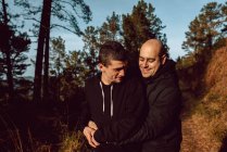 Cheerful homosexual couple embracing on walkway in forest in sunny day on blurred background — Stock Photo