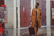 African American elegant woman with baggage walking on street near small shops — Stock Photo
