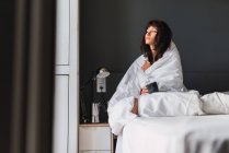 Молода щаслива жінка з чашкою напою, вкрита ковдрою, сидить на ліжку в спальні — стокове фото