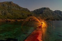 Long exposure of silhouette of human with burning round and fireworks on pier near water and mountains in evening — Stock Photo