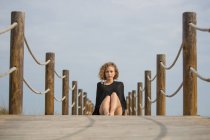 Молодая серьезная женщина сидит на деревянном пешеходном мосту и смотрит в камеру — стоковое фото