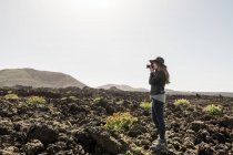 Vista lateral da fêmea em roupa casual usando câmera fotográfica para tirar fotos de terreno pedregoso maravilhoso no dia ensolarado — Fotografia de Stock