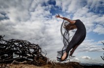 De baixo da bailarina jovem no desgaste preto com as mãos levantadas no ar perto de ramos secos e céu azul em nuvens — Fotografia de Stock