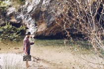 Вид сбоку на путешествующего человека, который держит небольшой чехол и пользуется телефоном, стоя на песчаном берегу спокойного озера против скалы — стоковое фото