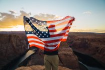 Vista posteriore dell'uomo con la bandiera sventolante USA in piedi vicino a un bellissimo canyon contro il cielo del tramonto sulla costa occidentale — Foto stock