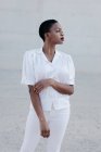 Елегантна короткошерста етнічна жінка в білій сорочці і штанях позує на сіру стіну — стокове фото