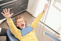 Heureux garçon couché dans la clinique dentaire en regardant la caméra avec les bras tendus — Photo de stock