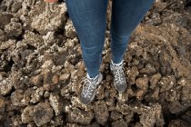 Сверху ноги анонимной женщины в джинсах и кроссовках стоят на скалистой земле в сельской местности — стоковое фото