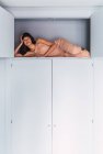 Portrait of young woman lying on high shelf of wardrobe in bedroom — Stock Photo