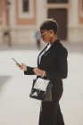 Уверенная афроамериканская элегантная женщина в костюмах и солнцезащитных очках держит сумку и мобильный телефон на улице — стоковое фото